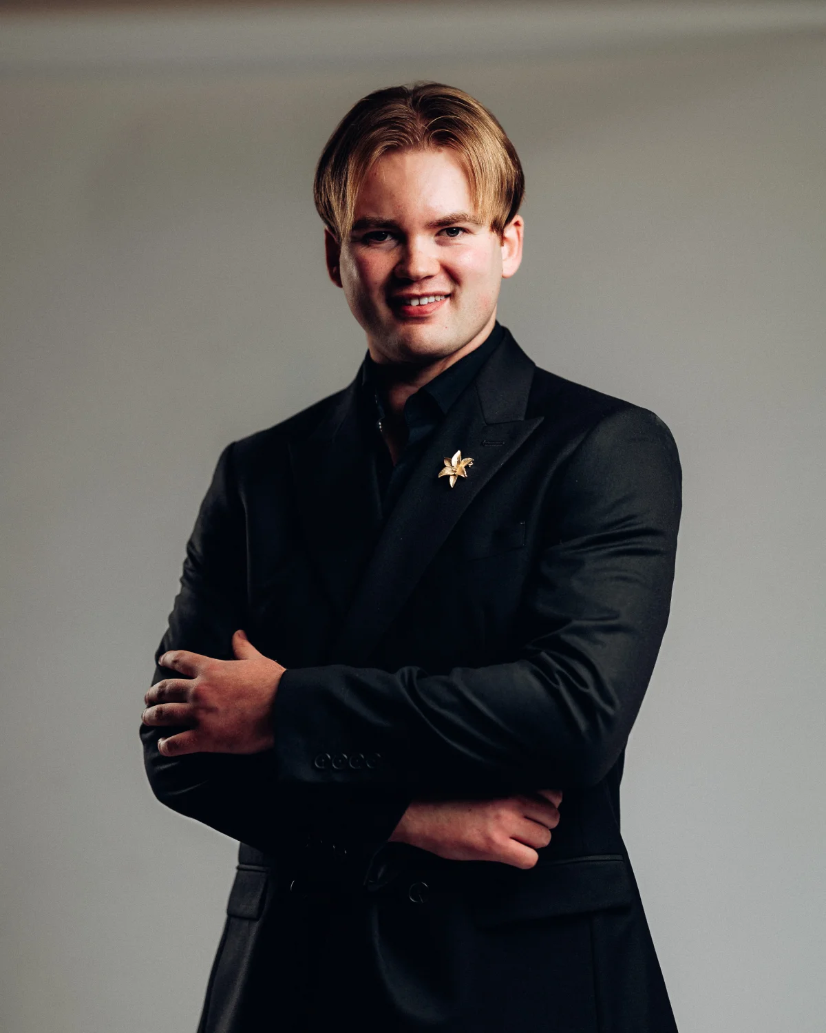Professional portrait of a young man in a black suit with crossed arms against a neutral grey background.