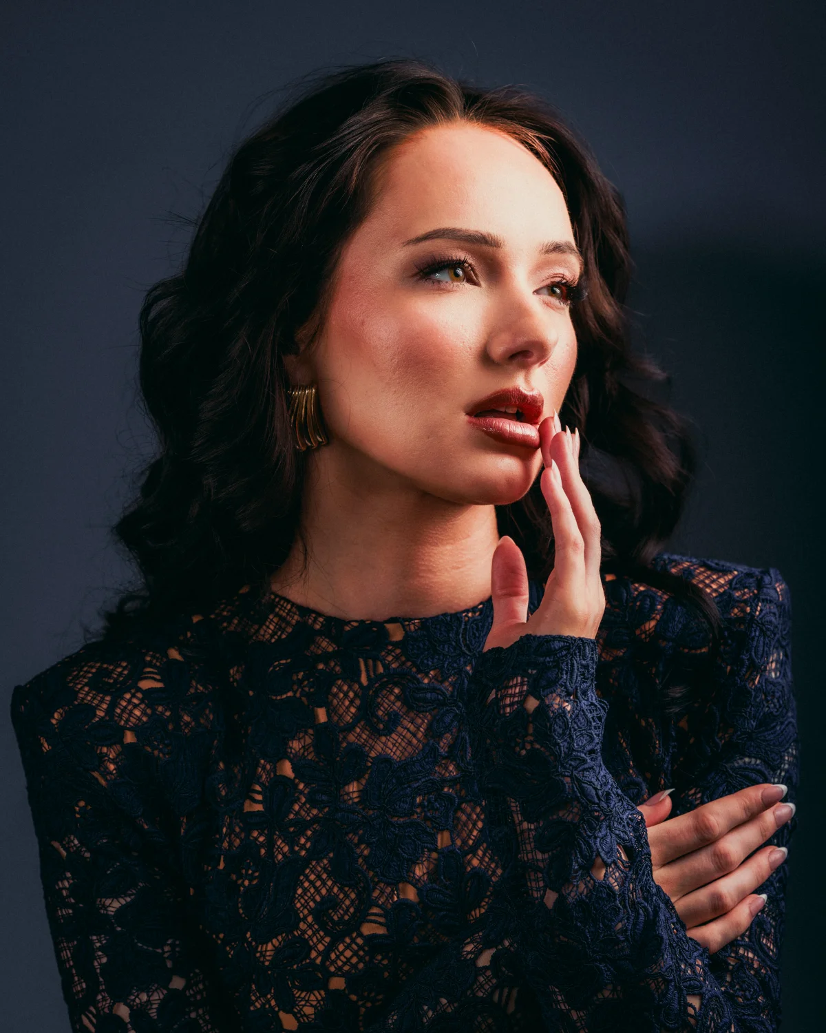 Close-up portrait of a woman with dark wavy hair and red lipstick in a navy blue lace dress.