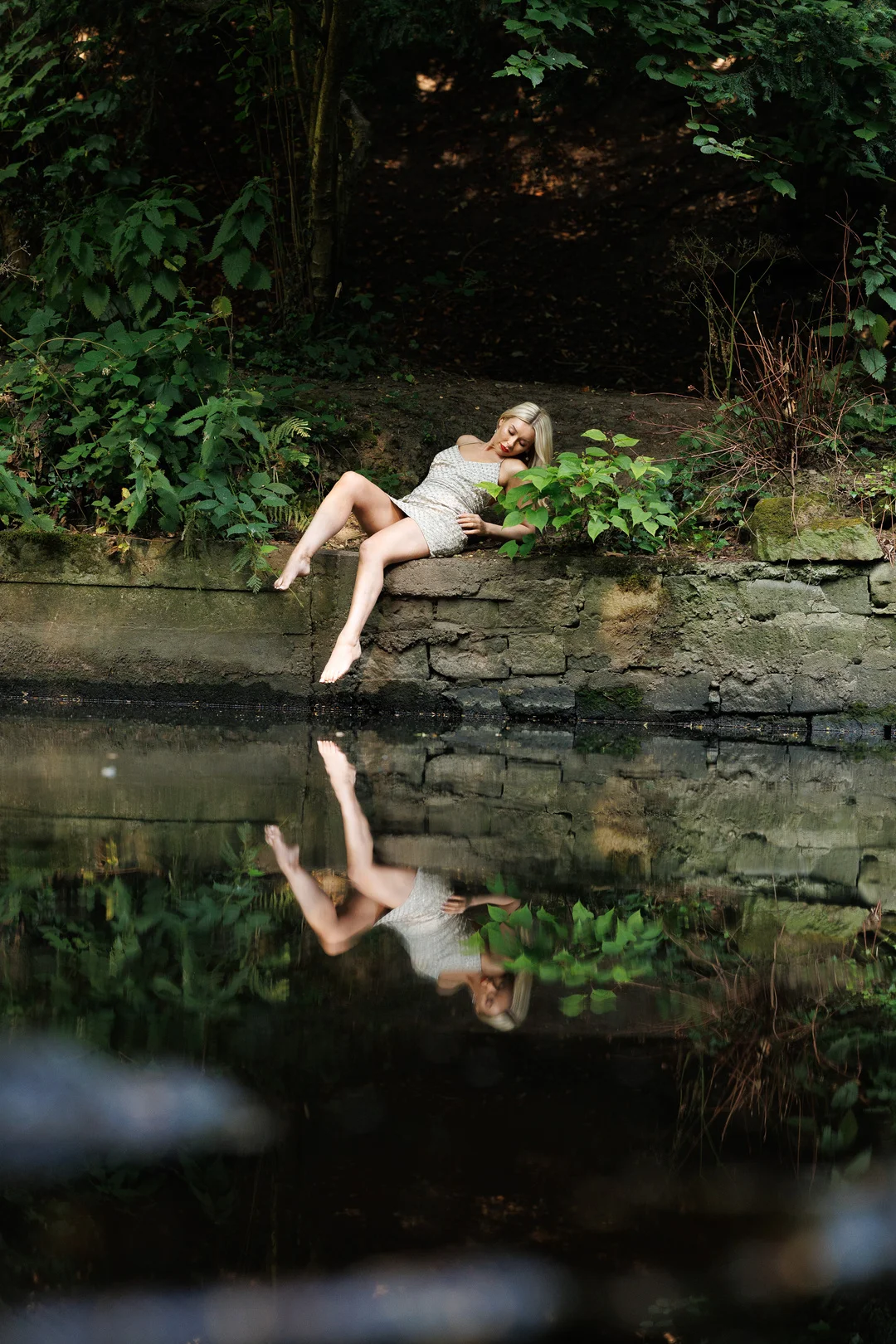 A woman in a short white dress reclining on a stone wall by a pond, with her reflection in the water.