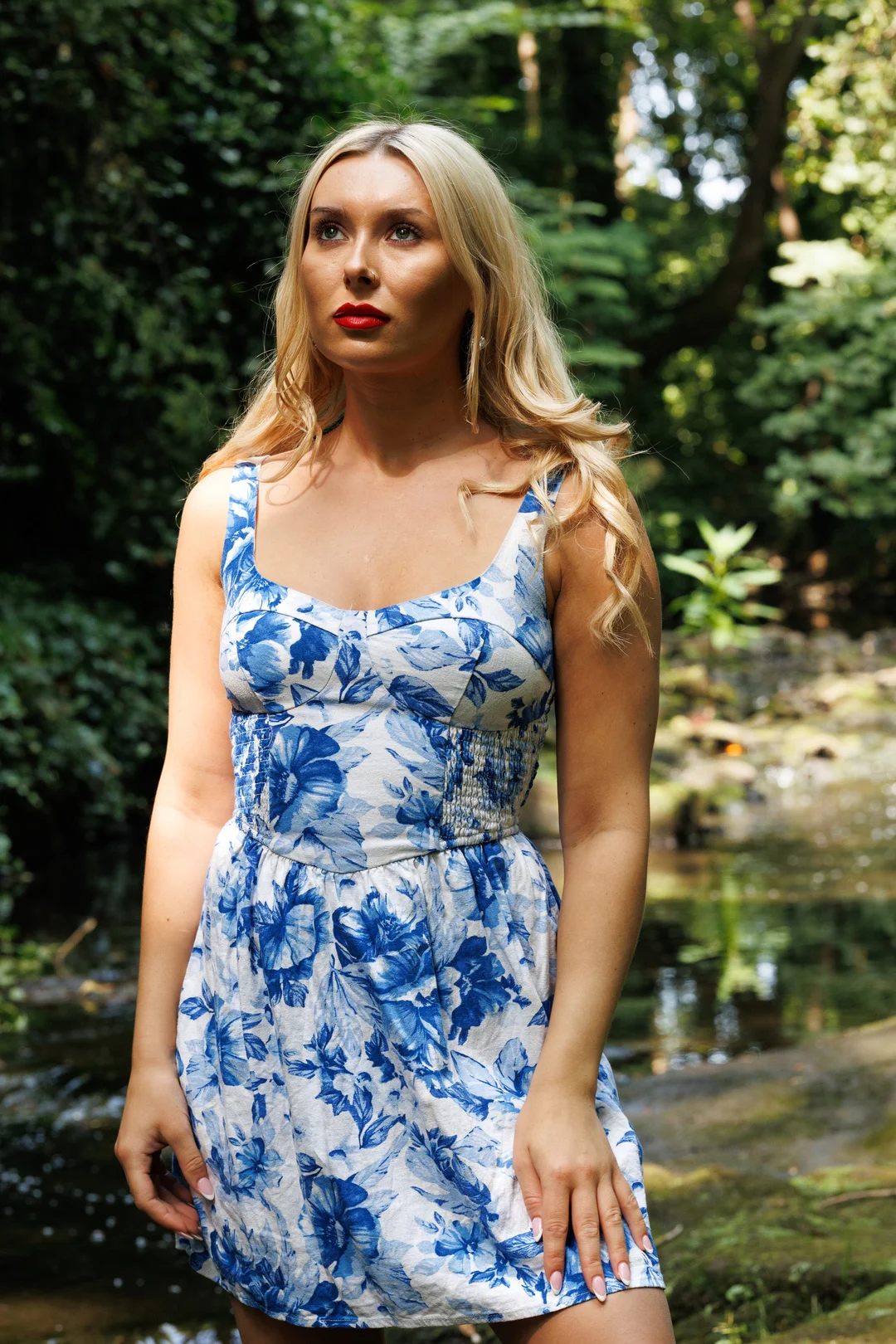 Blonde woman in a blue and white floral dress posing in a lush green forest by a stream.
