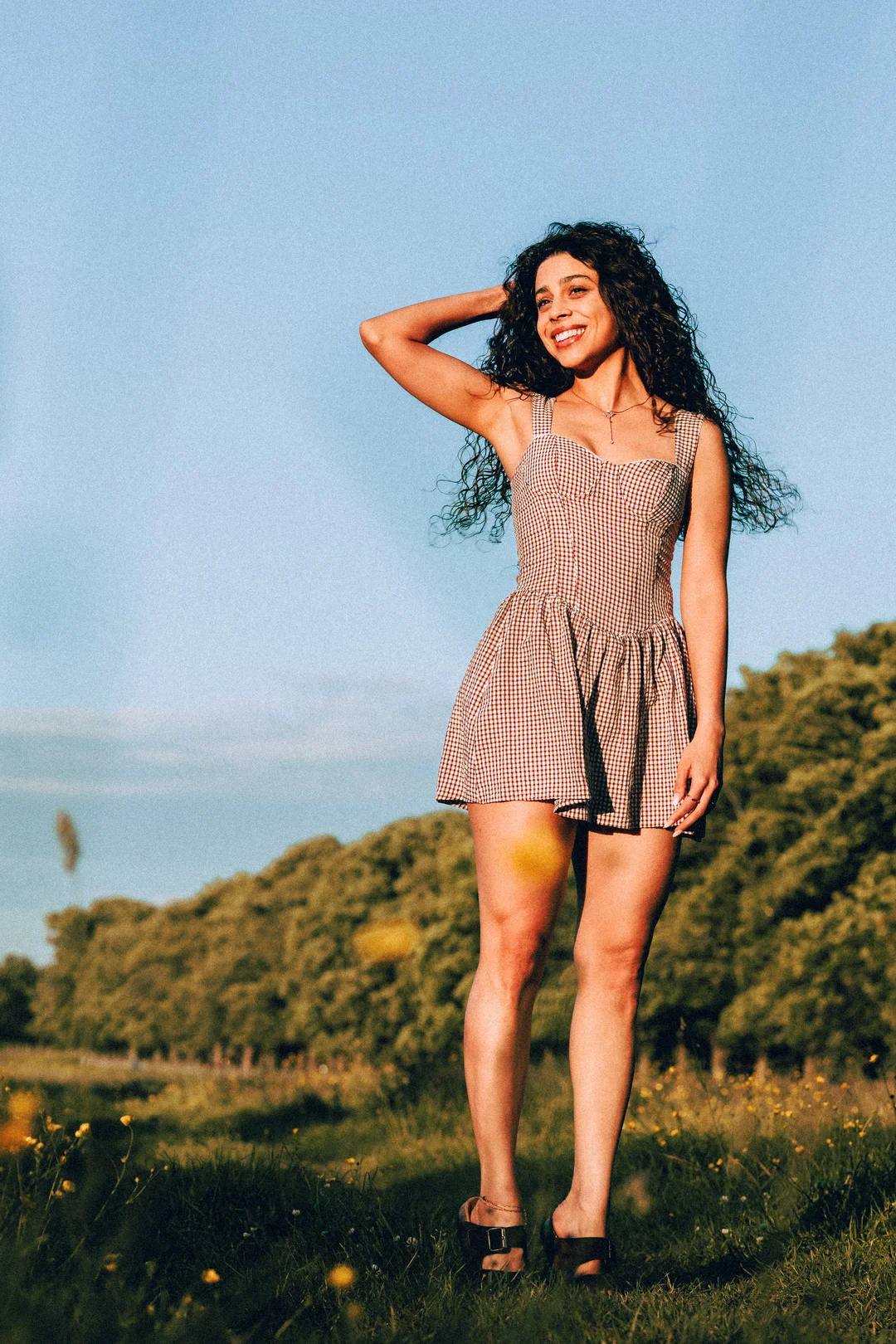 Smiling woman with curly hair in a red gingham dress posing in a sunny meadow.