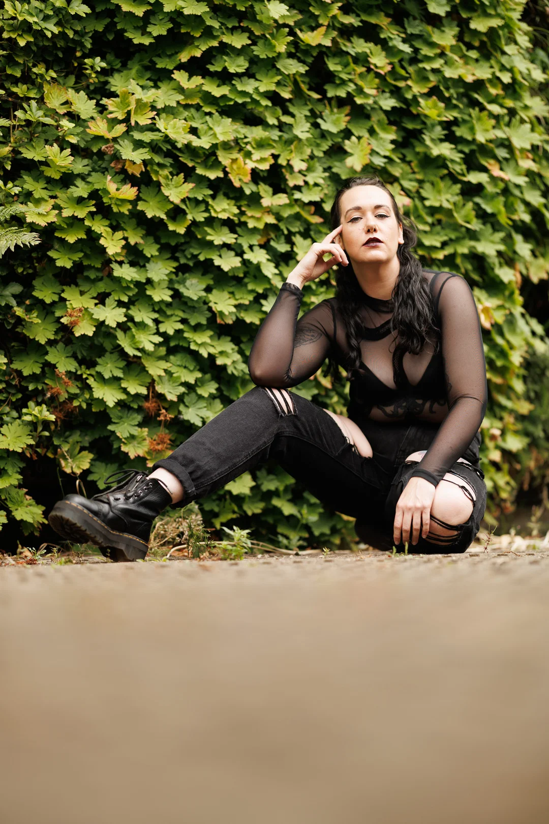 Woman with dark makeup and a black mesh top posing in front of a lush green hedge.