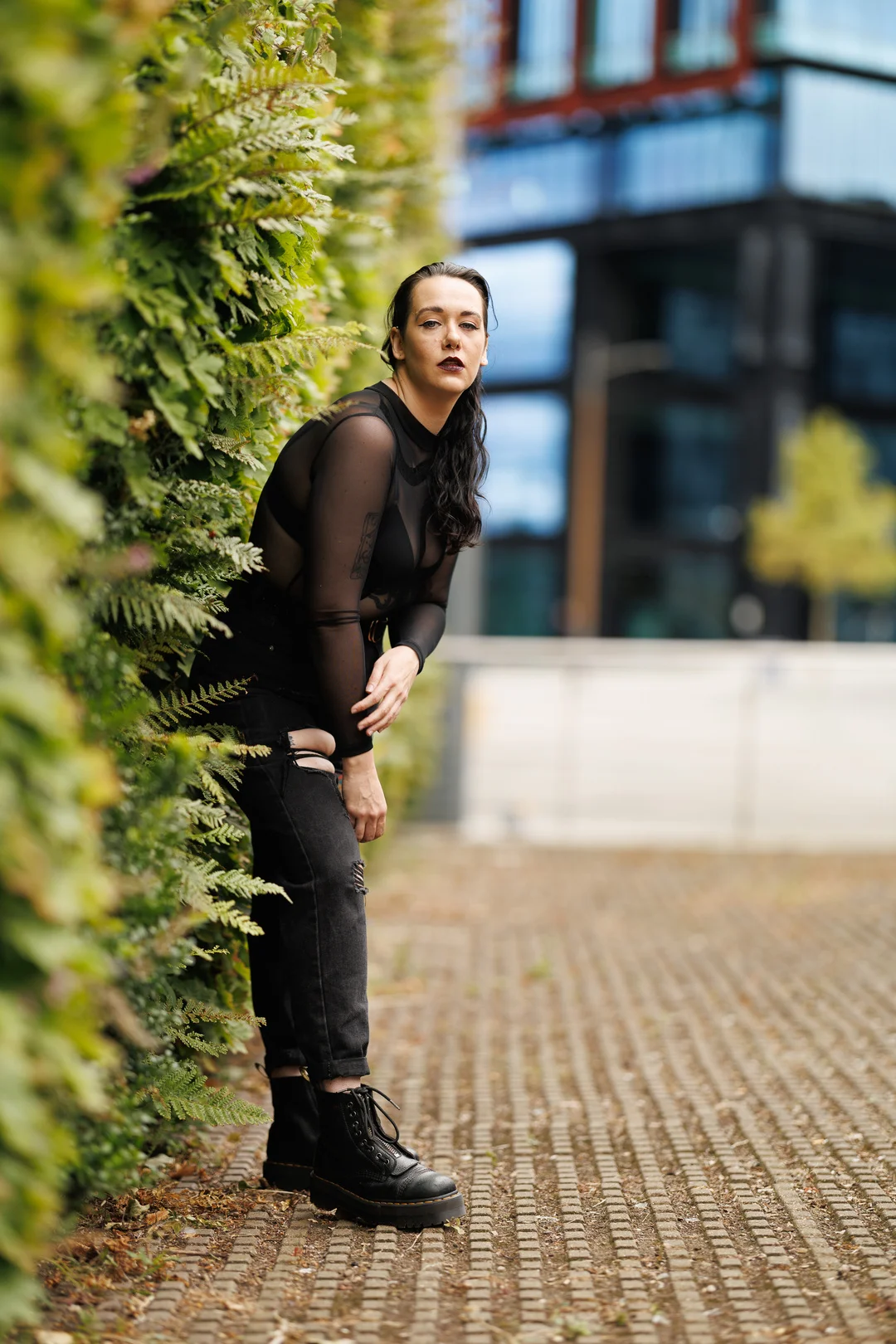 A person in a black sheer top and ripped jeans posing against a lush green hedge in an urban environment.