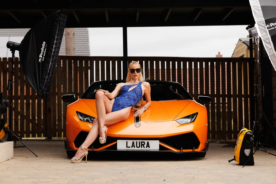 Woman in a revealing blue dress posing on an orange Lamborghini with studio lighting equipment visible.