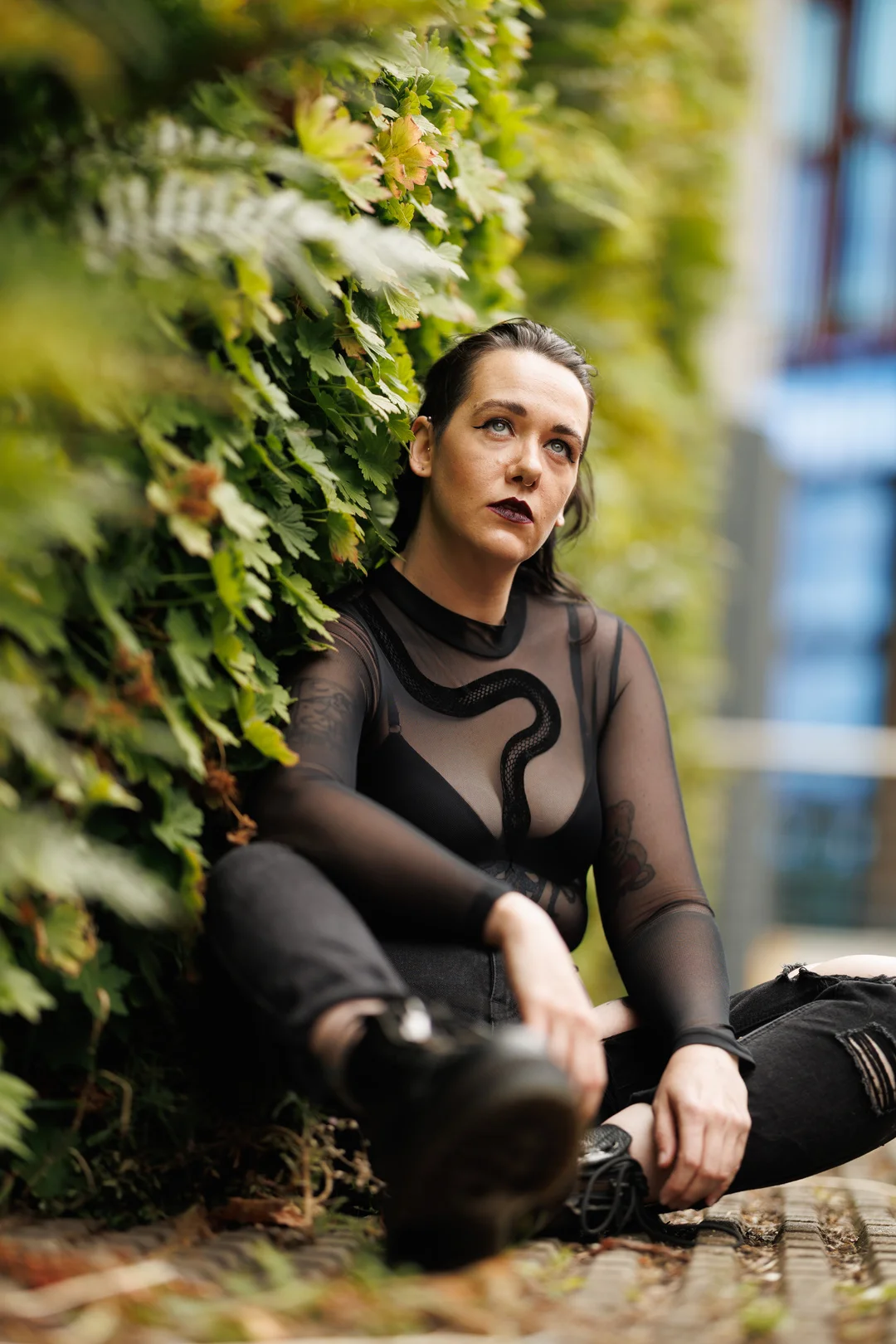 A person with dark makeup and a black mesh top sitting against a lush green hedge.