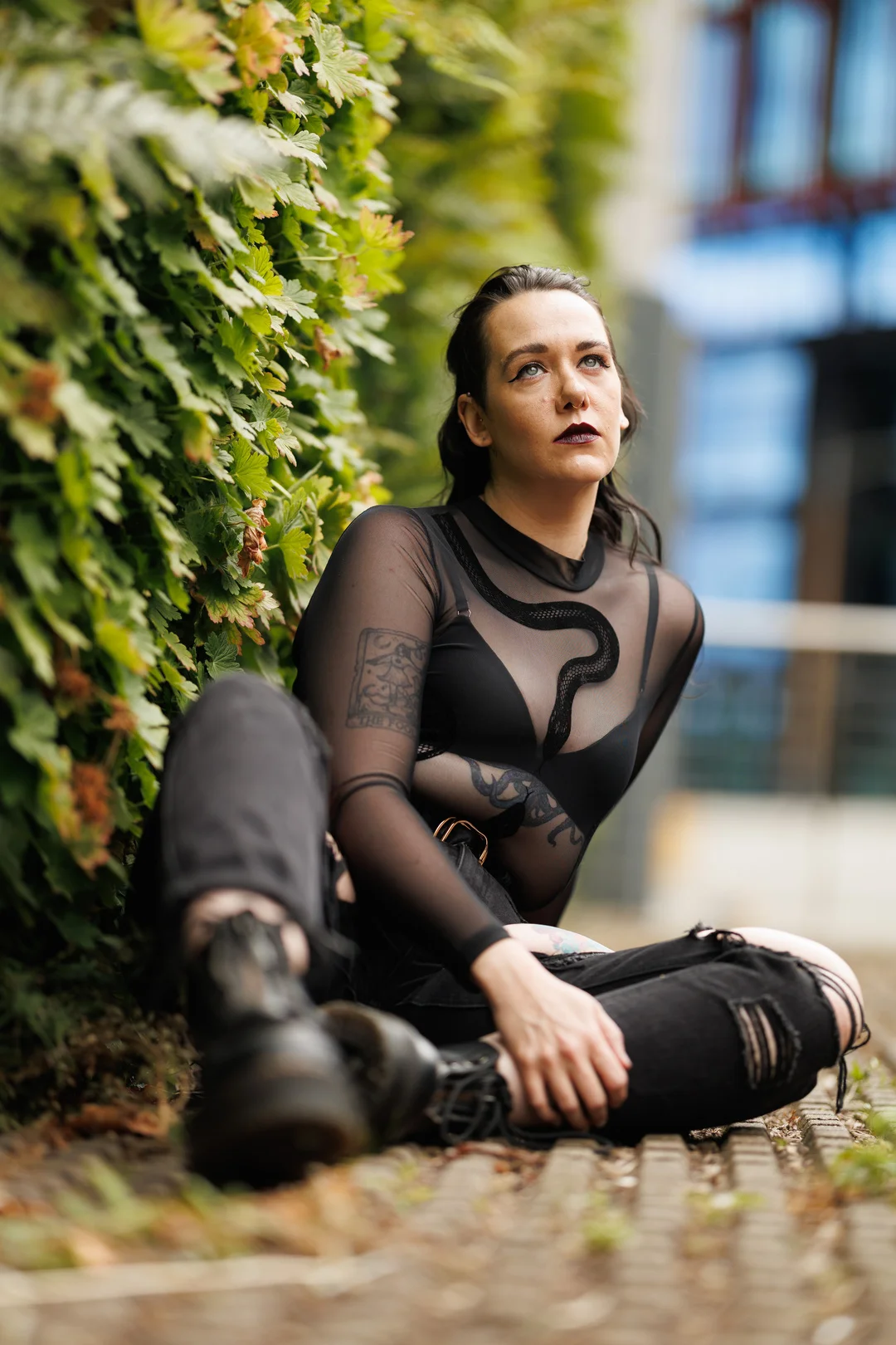 A woman with dark lipstick and a black sheer top sitting outdoors against a green hedge.