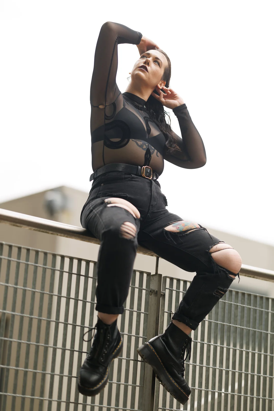 Woman in a sheer black top and ripped black jeans posing on a metal railing against a bright sky.