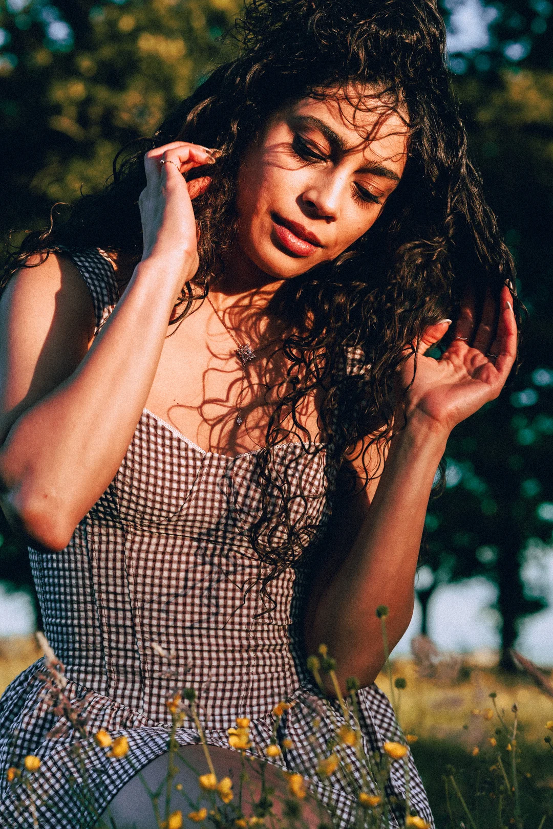 Woman with curly dark hair and a checkered dress posing in a sunlit field.