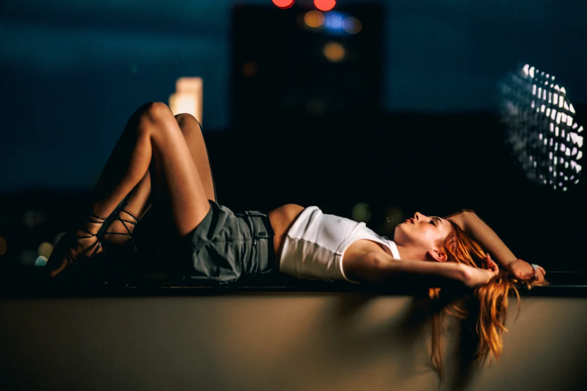 A woman with red hair lying on a ledge at night, wearing a white crop top and grey skirt, in a moody urban setting.