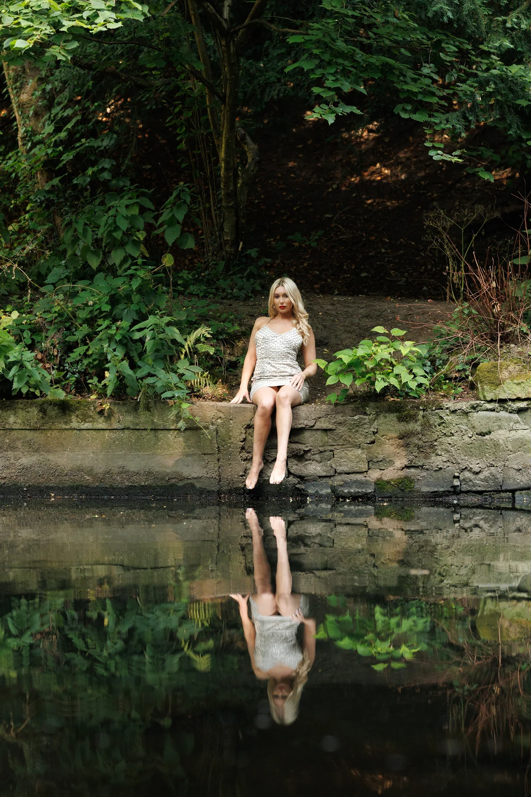 Woman in a sparkly silver dress sitting on a stone wall by a pond, reflecting in the still water.