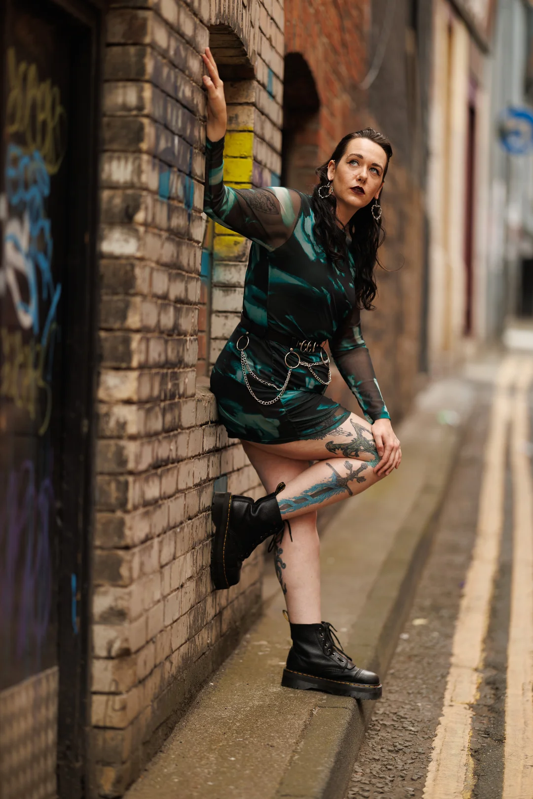 Woman with tattoos and dark makeup posing in a green dress and black boots against an urban brick wall.