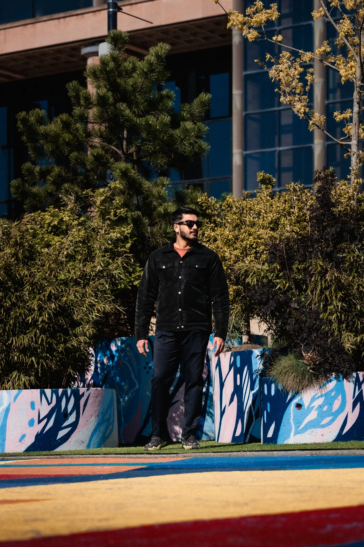 Full-body shot of a man in a black jacket and trousers posing outdoors near colorful art and greenery.