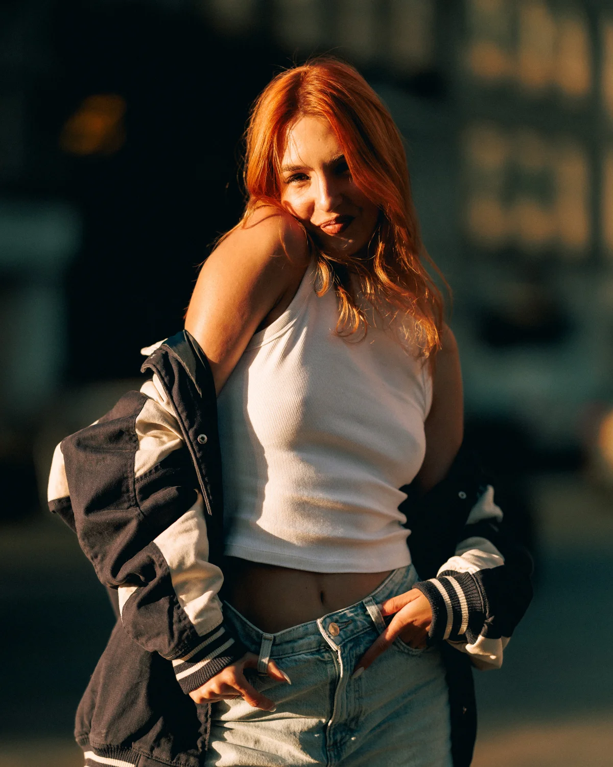 A young woman with red hair in a white tank top and varsity jacket posing in warm outdoor sunlight.