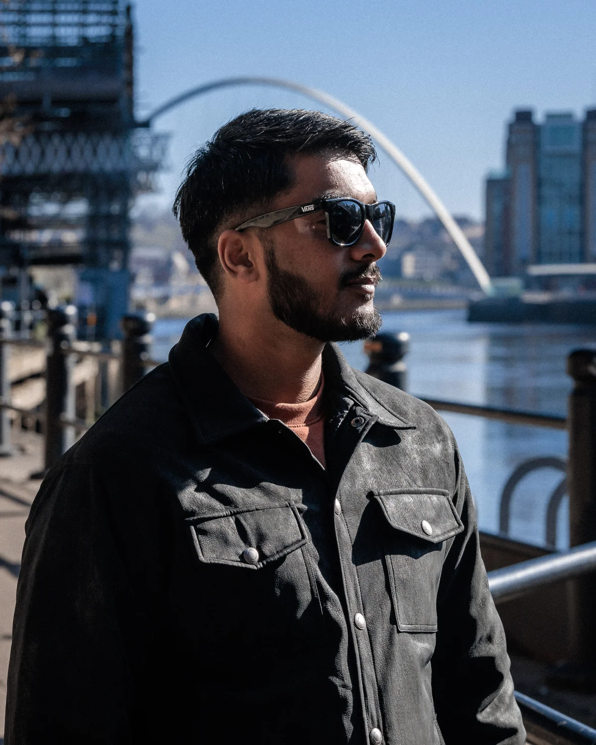 Side profile of a bearded man in a black jacket and sunglasses against an urban waterfront backdrop.