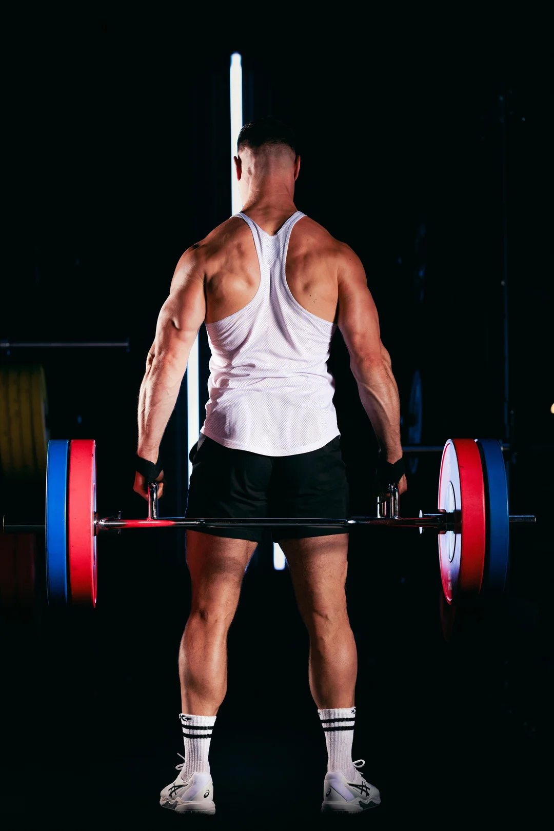 Muscular man seen from behind holding a barbell in a dark gym, emphasizing athletic physique.