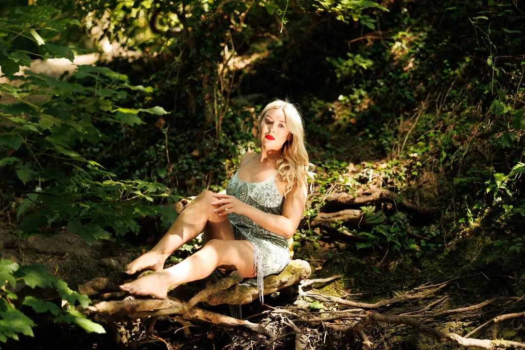 Blonde woman in a light-colored dress posing on a log in a lush green forest.
