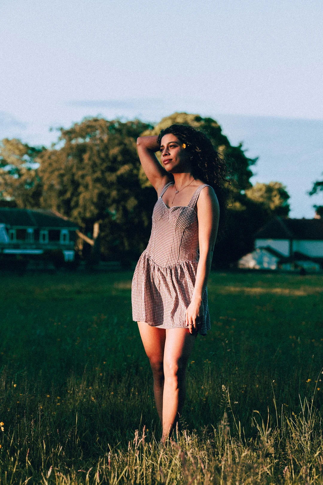 Woman with curly hair in a red gingham dress posing in a sunlit grassy field.