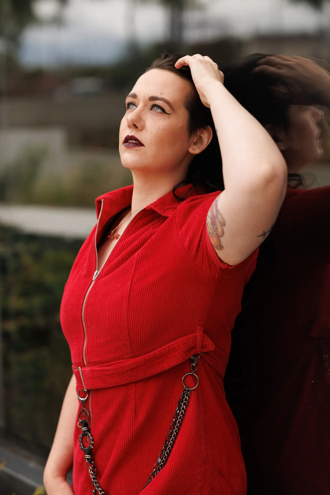 A person in a red corduroy dress with chains, leaning against a reflective surface, looking upwards.