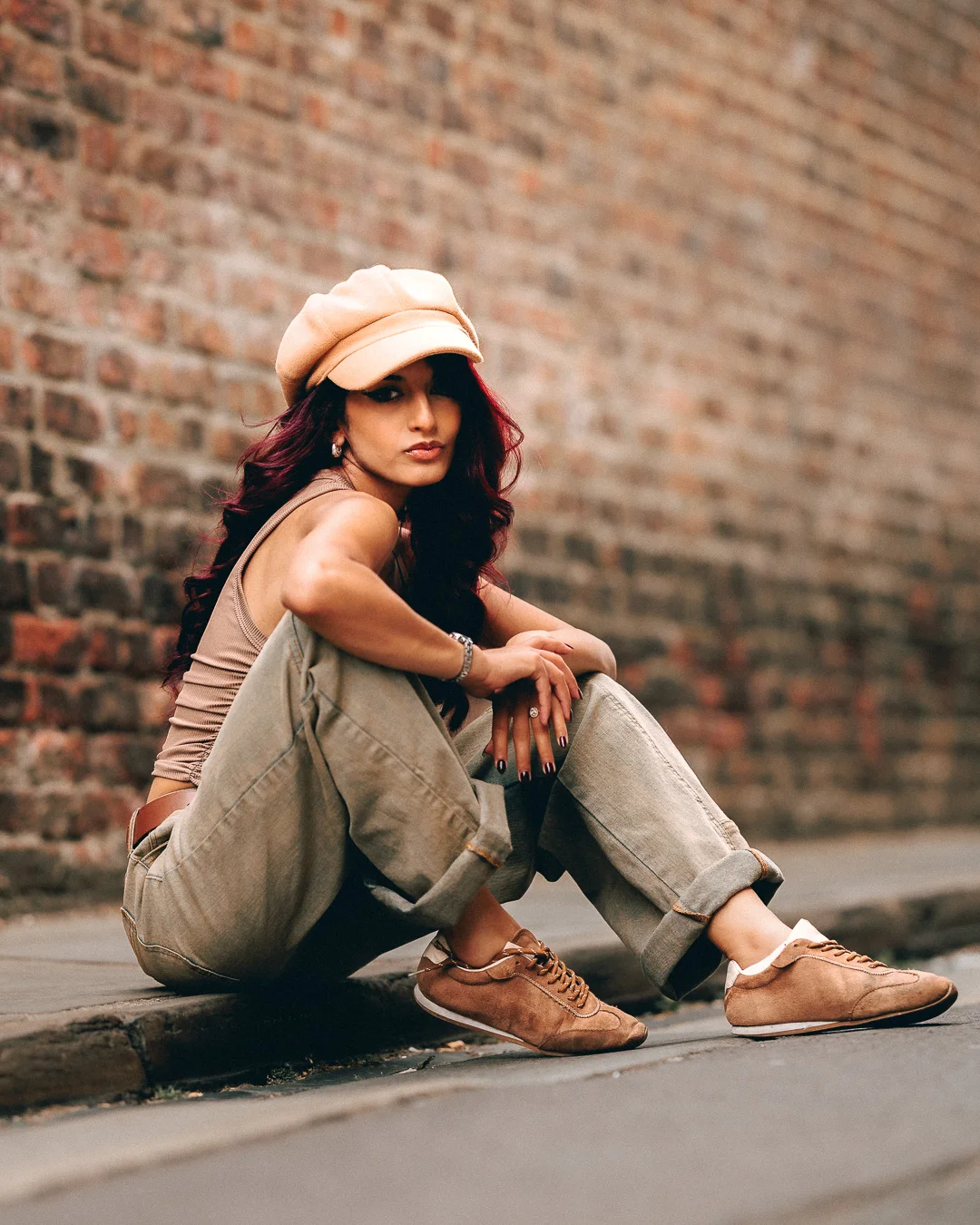 Woman with red hair in a beige cap and khaki outfit sitting against a brick wall in an urban setting.
