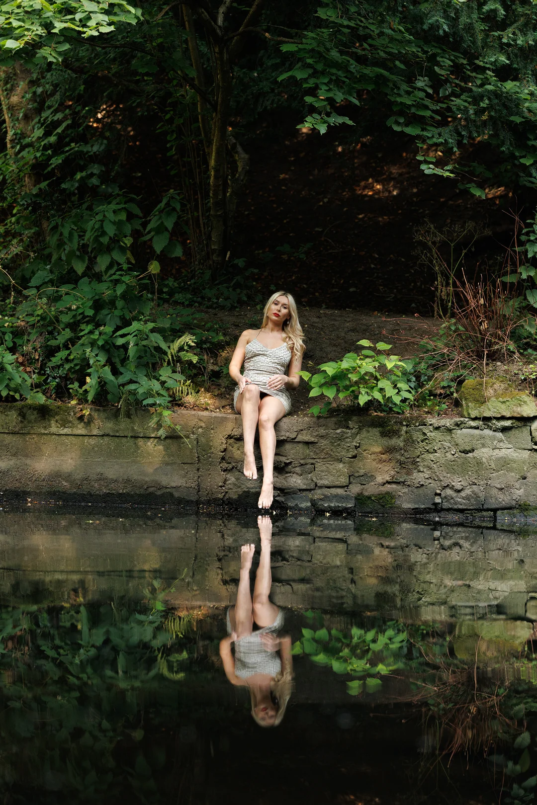 Blonde woman in a short dress sitting on a stone ledge by a pond, reflecting in the still water.