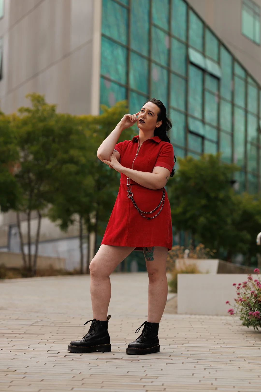 A person in a short red zip-up dress and black combat boots posing in front of a modern glass building.