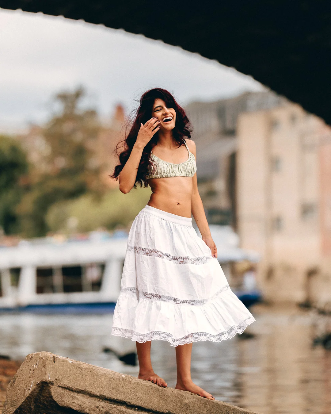 A laughing woman with long dark hair in a crochet top and white skirt standing on a rock by a river.