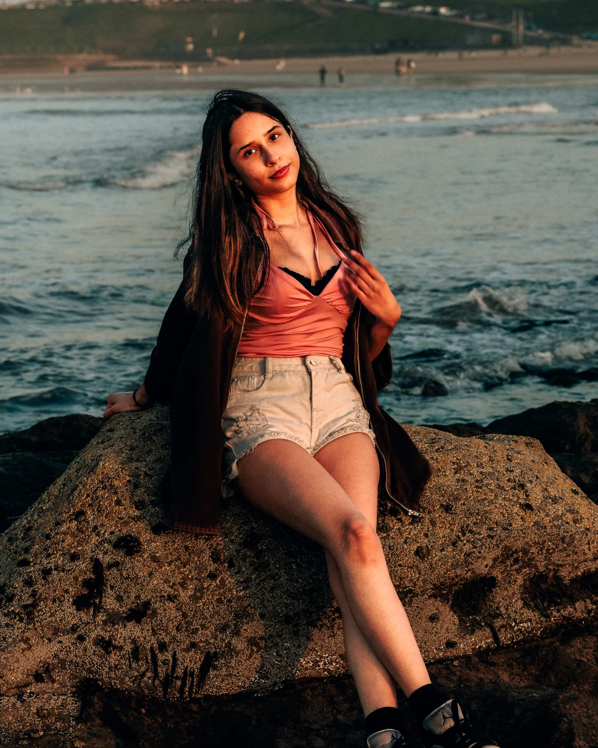Young woman with long dark hair sitting on a rock at a beach during golden hour, wearing a pink top and white shorts.