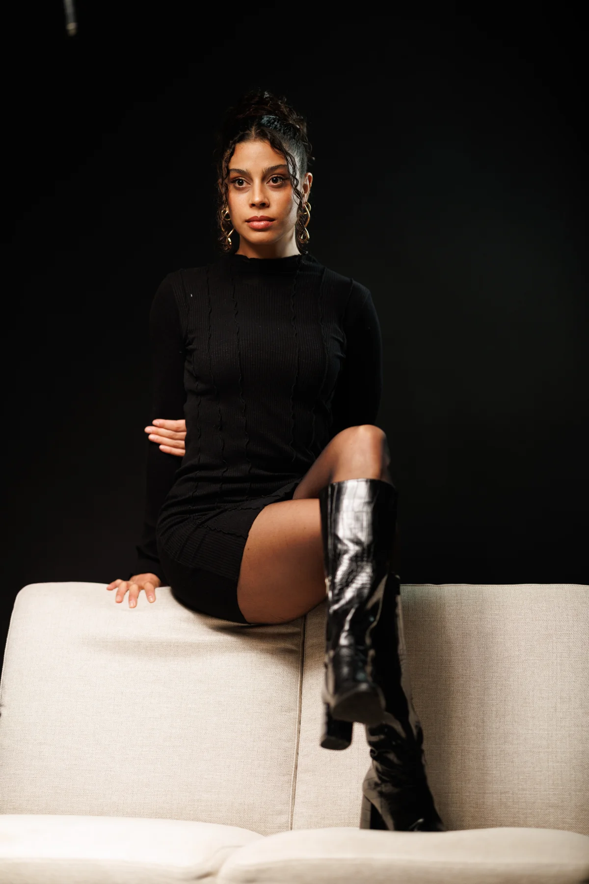 Woman in a black mini dress and tall black boots posing on a white couch against a black background.