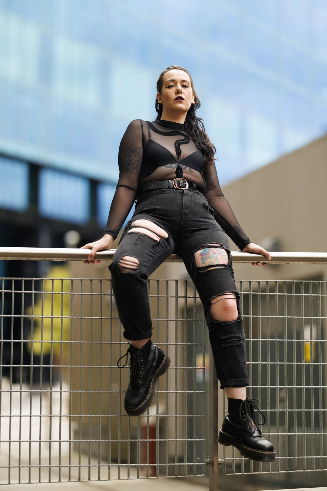 Woman in a sheer black top and ripped jeans posing on a metal railing in an urban setting.