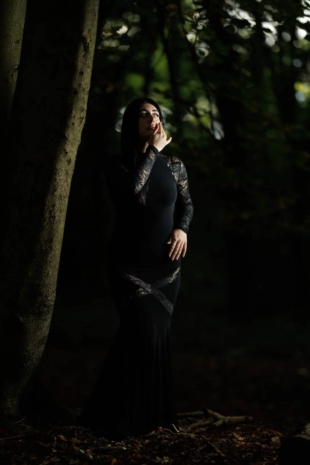 A woman in a long, form-fitting black lace dress posing in a dark, moody forest.