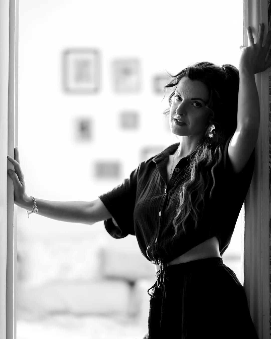 Black and white portrait of a woman with long wavy hair posing between two white walls.