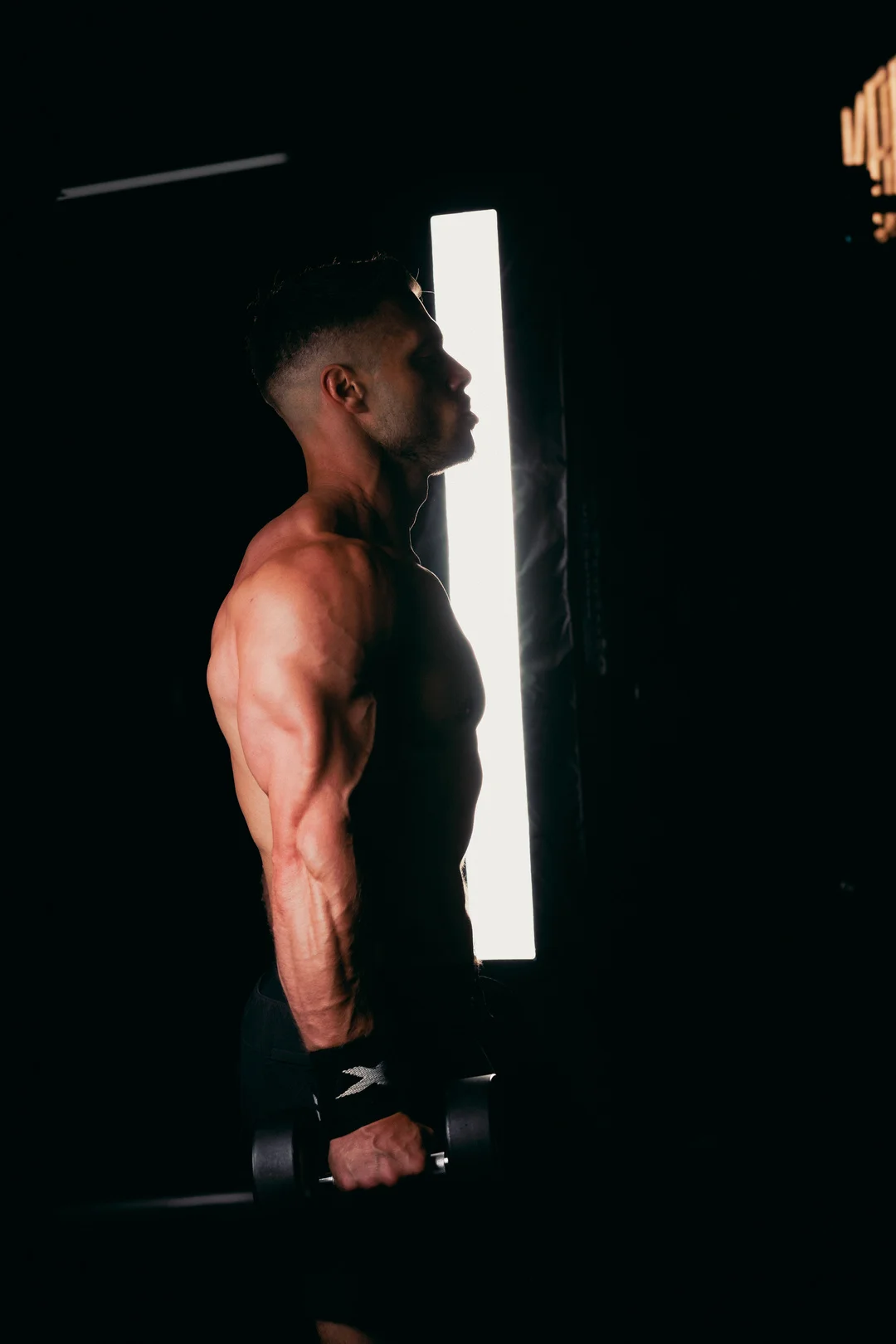 Side profile of a muscular shirtless man holding a dumbbell in a dark gym with dramatic side lighting.