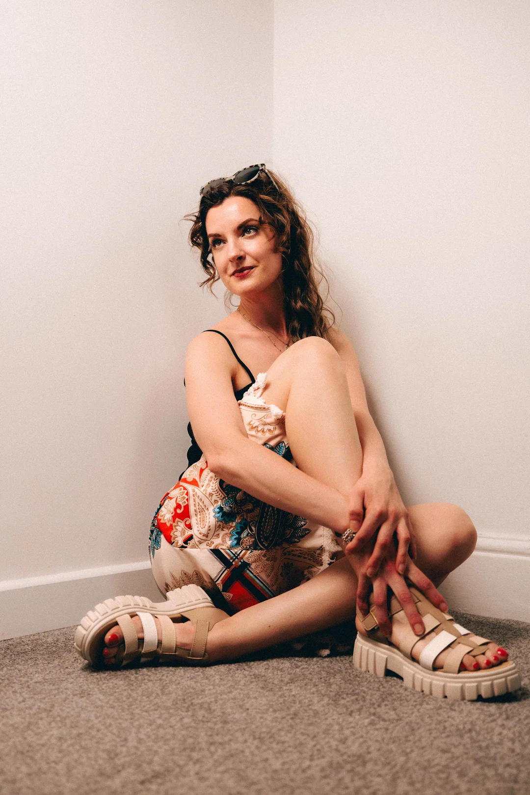 Woman with curly hair sitting on a carpeted floor in a black top, patterned skirt, and platform sandals.