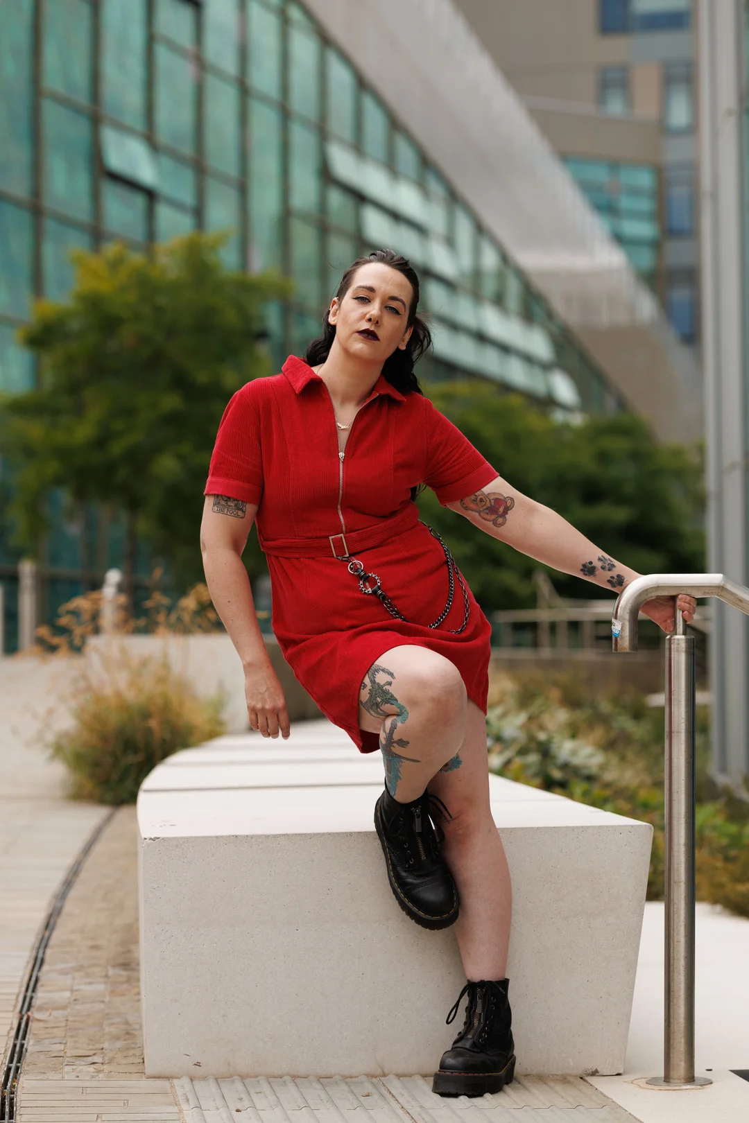 Woman with tattoos in a red dress and black boots posing on a white bench in an urban setting.