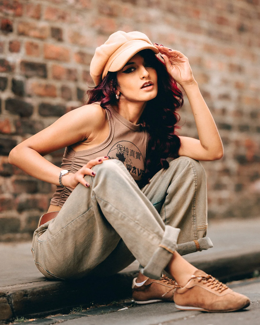 Woman with red hair in a tan cap and cargo pants posing against a brick wall.