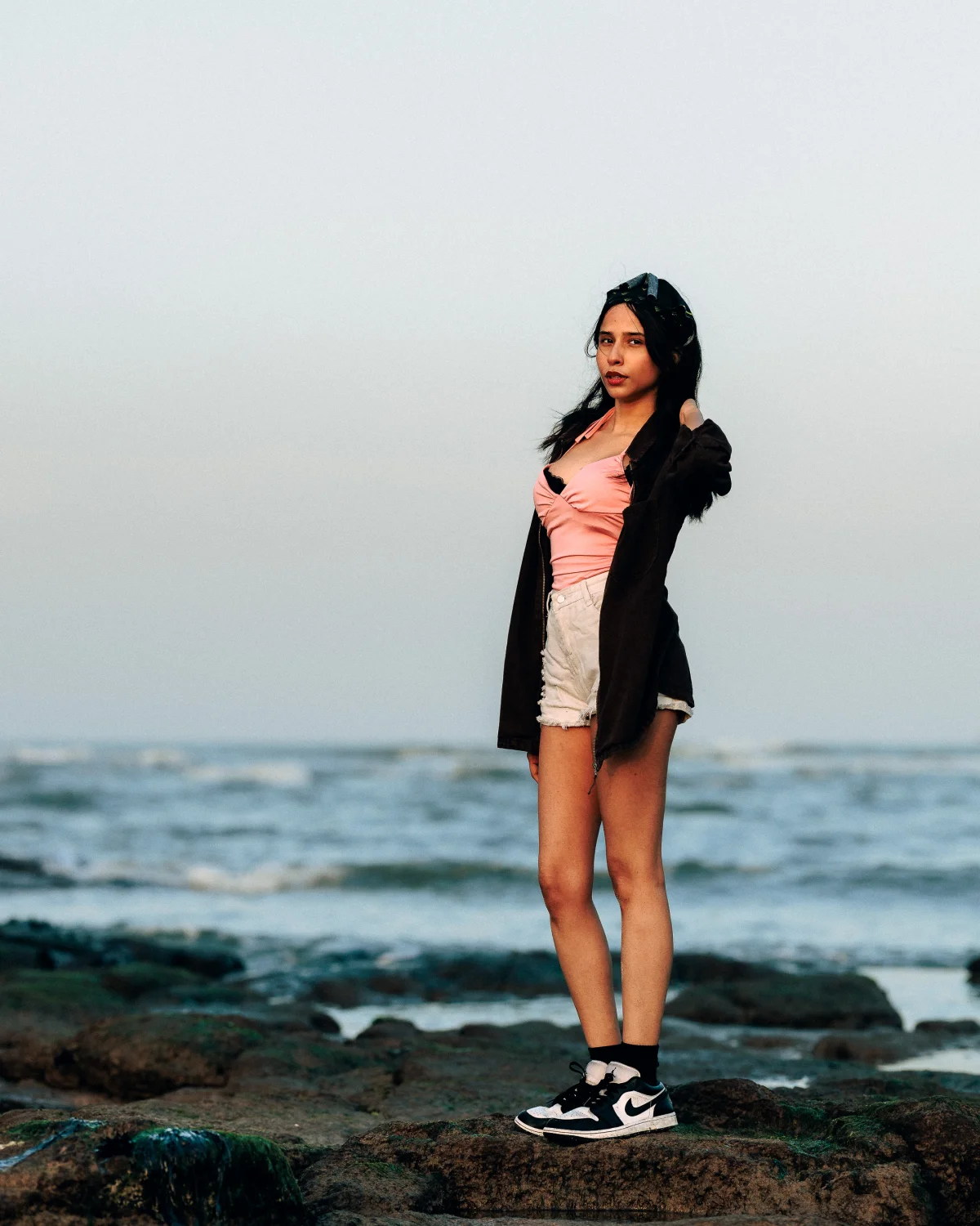 Young woman in a pink top and white shorts posing on a rocky beach against a cloudy sky and ocean.