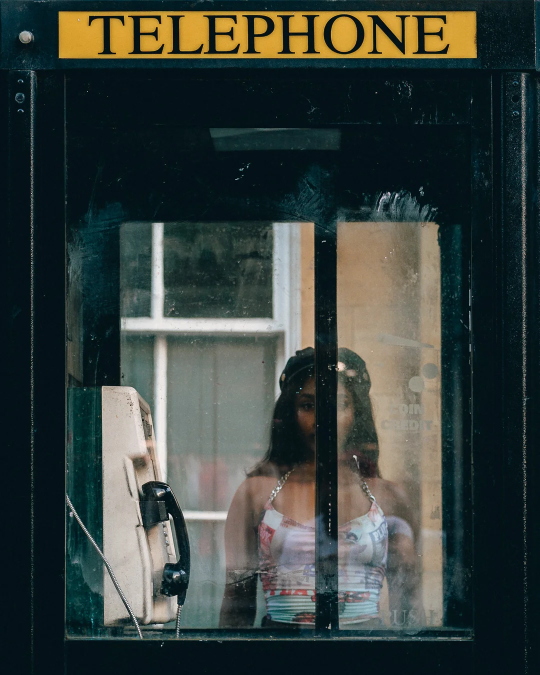 A woman standing inside a vintage telephone booth, viewed through reflective glass in a moody urban setting.