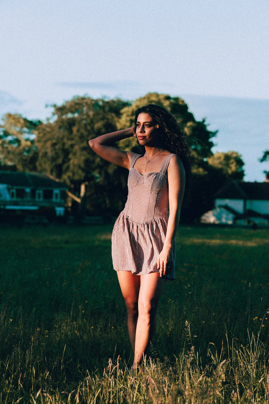 A woman with curly hair in a red and white gingham dress standing in a grassy field during golden hour.