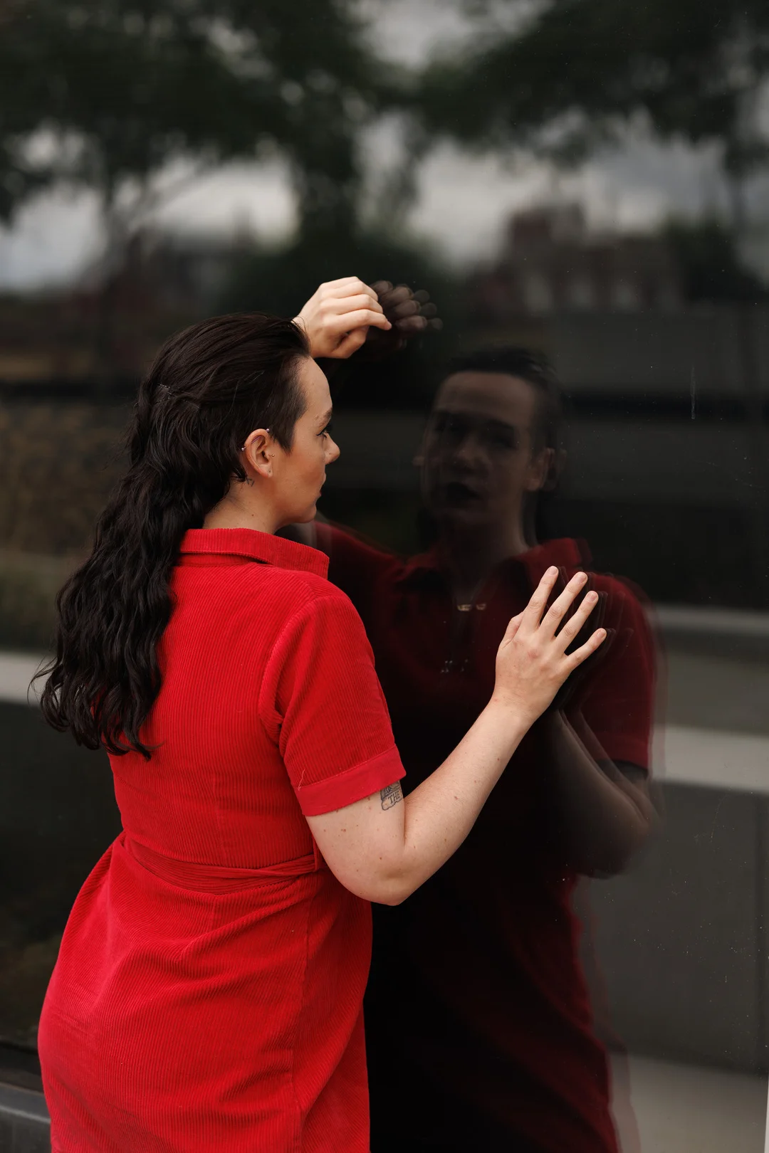 Woman in a red corduroy dress interacting with her reflection in a dark glass window.