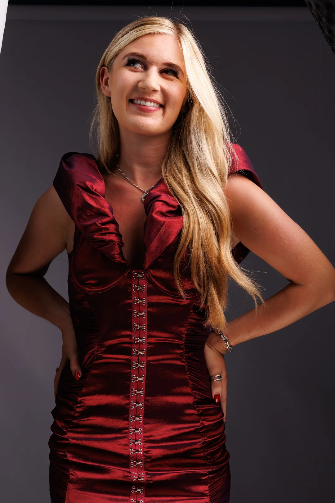 A smiling blonde woman in a red satin dress posing against a dark grey studio background.