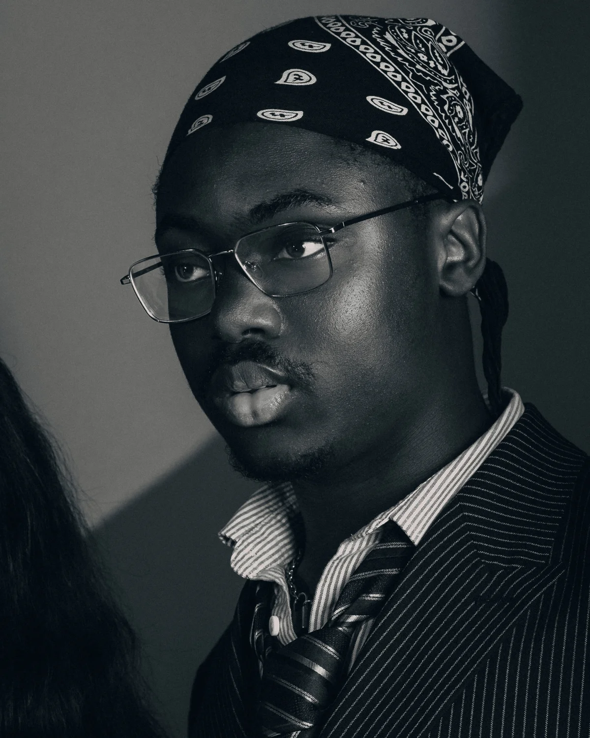 Black and white portrait of a Black man wearing glasses, a bandana, and a pinstripe suit jacket.