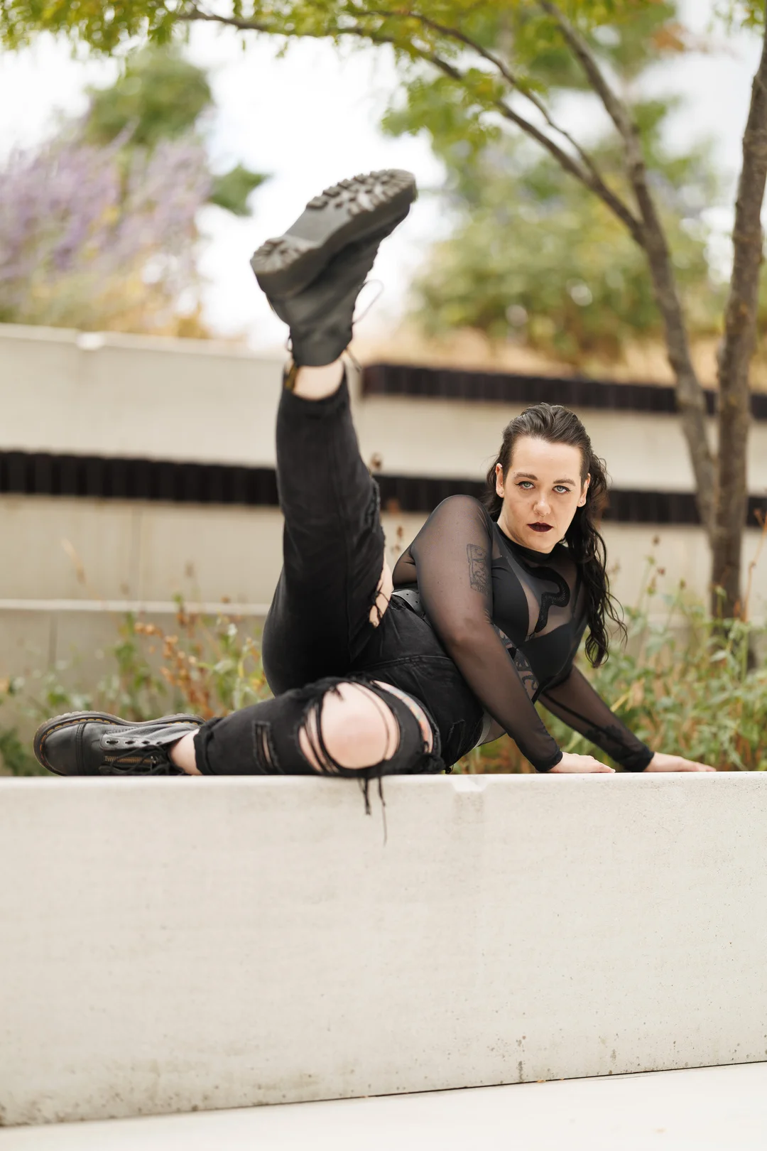 Woman in black mesh top and ripped jeans posing dynamically on a concrete ledge outdoors.