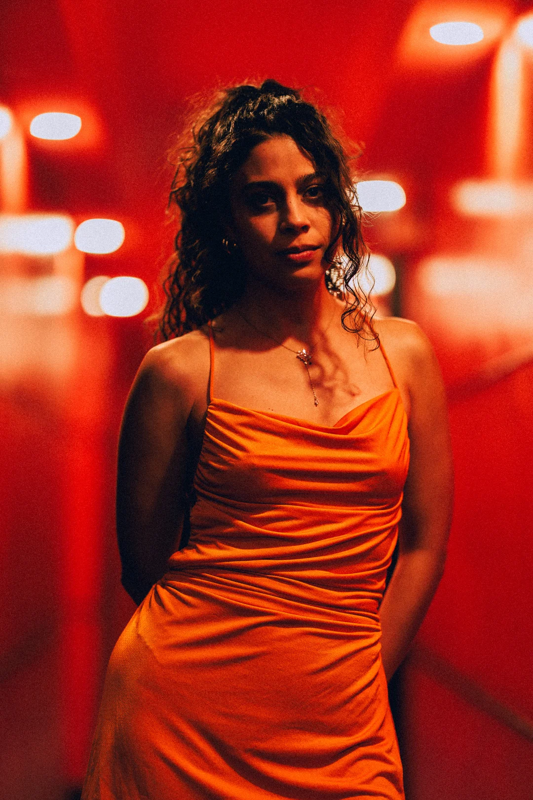 Woman with curly hair in an orange satin dress against a moody red background with bokeh lights.