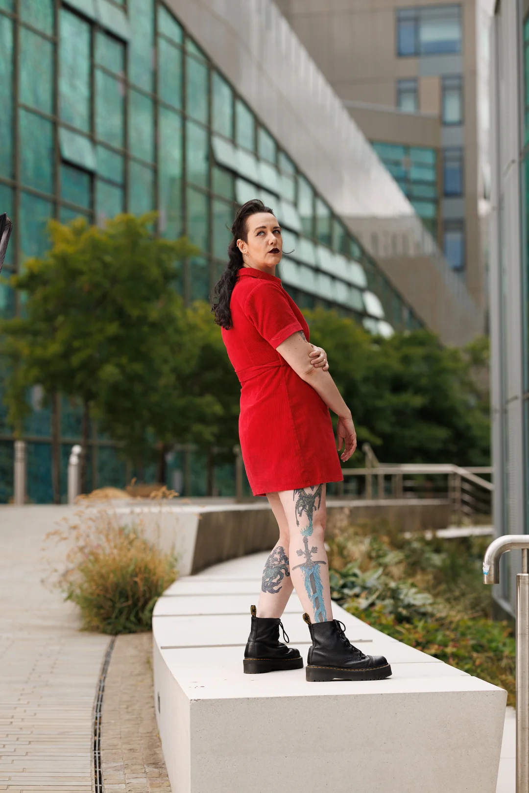 Woman in a red dress and black boots posing against a modern glass building background.