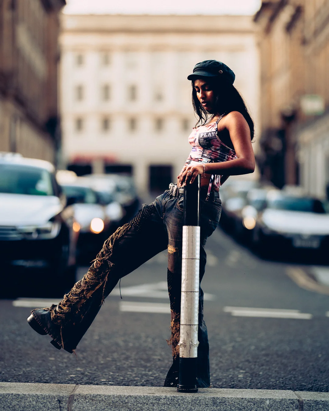 A woman in a fashion-forward outfit with distressed jeans and a cap posing against a bollard on a city street.