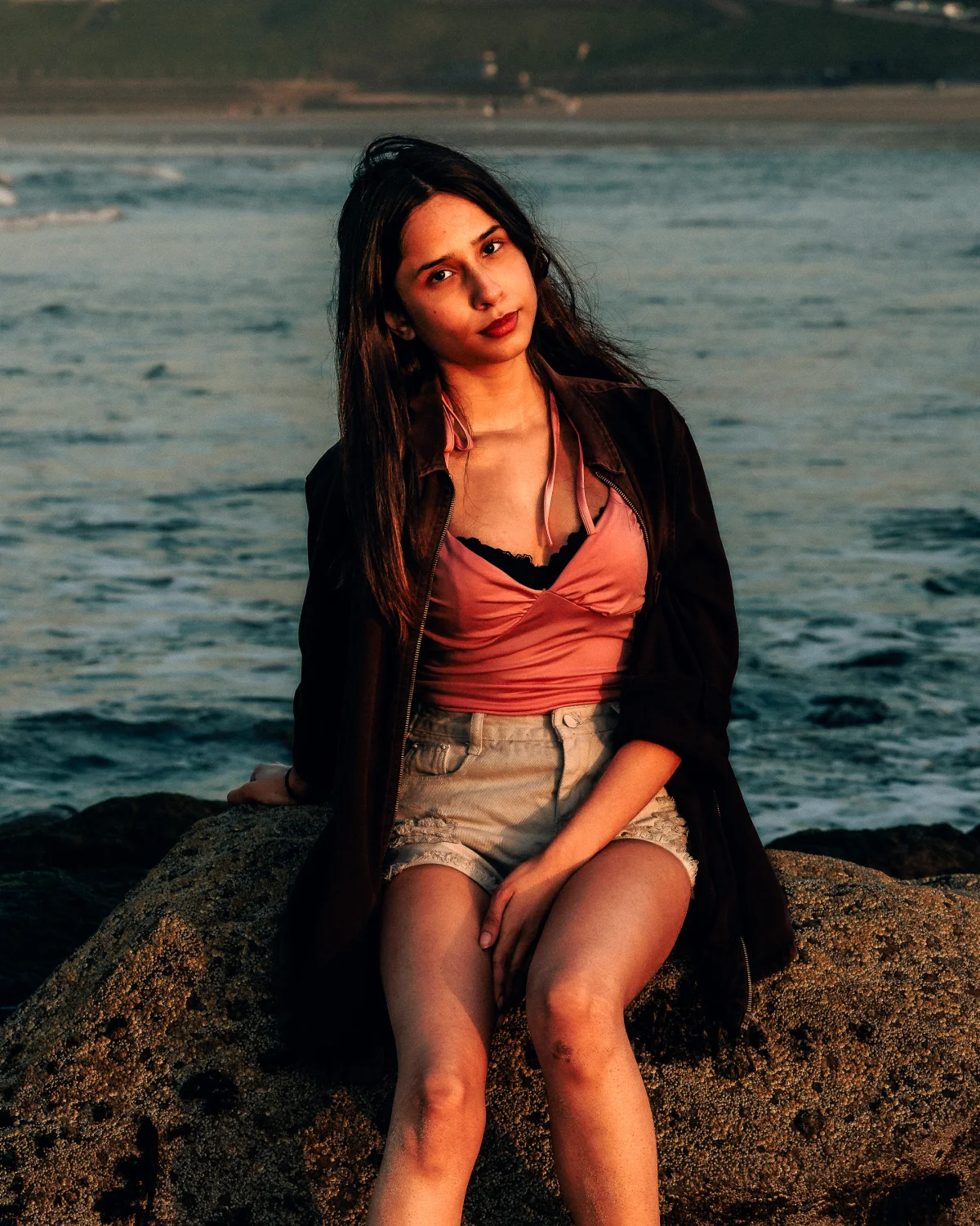 Young woman with long dark hair posing on a rock by the ocean at sunset, wearing a pink top and white shorts.