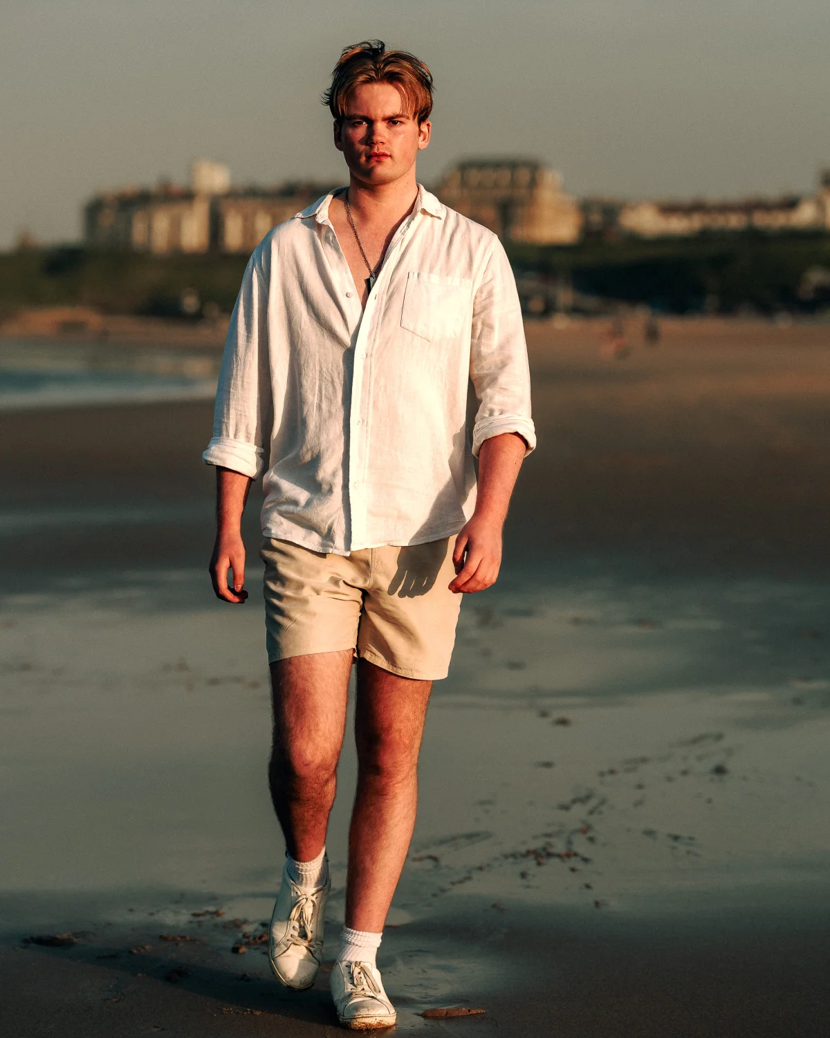 A young man in a white linen shirt and beige shorts walking on a beach during golden hour.