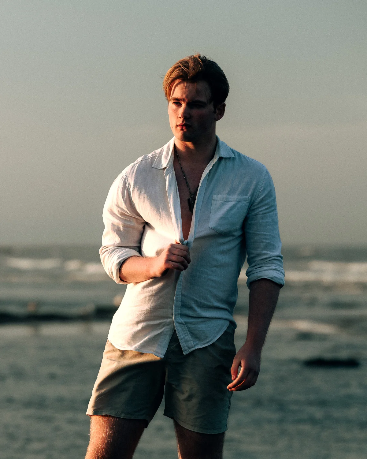 A man in a white linen shirt and khaki shorts posing on a beach during golden hour.