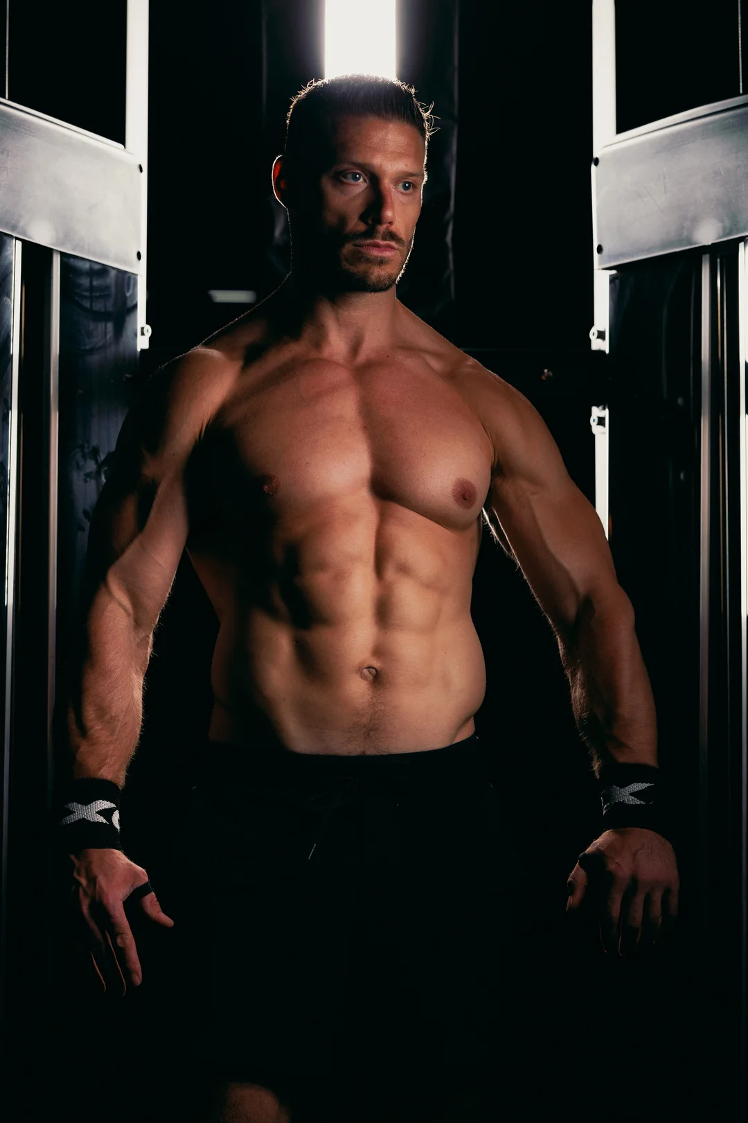 Muscular shirtless man standing in a gym with dramatic low-key lighting.