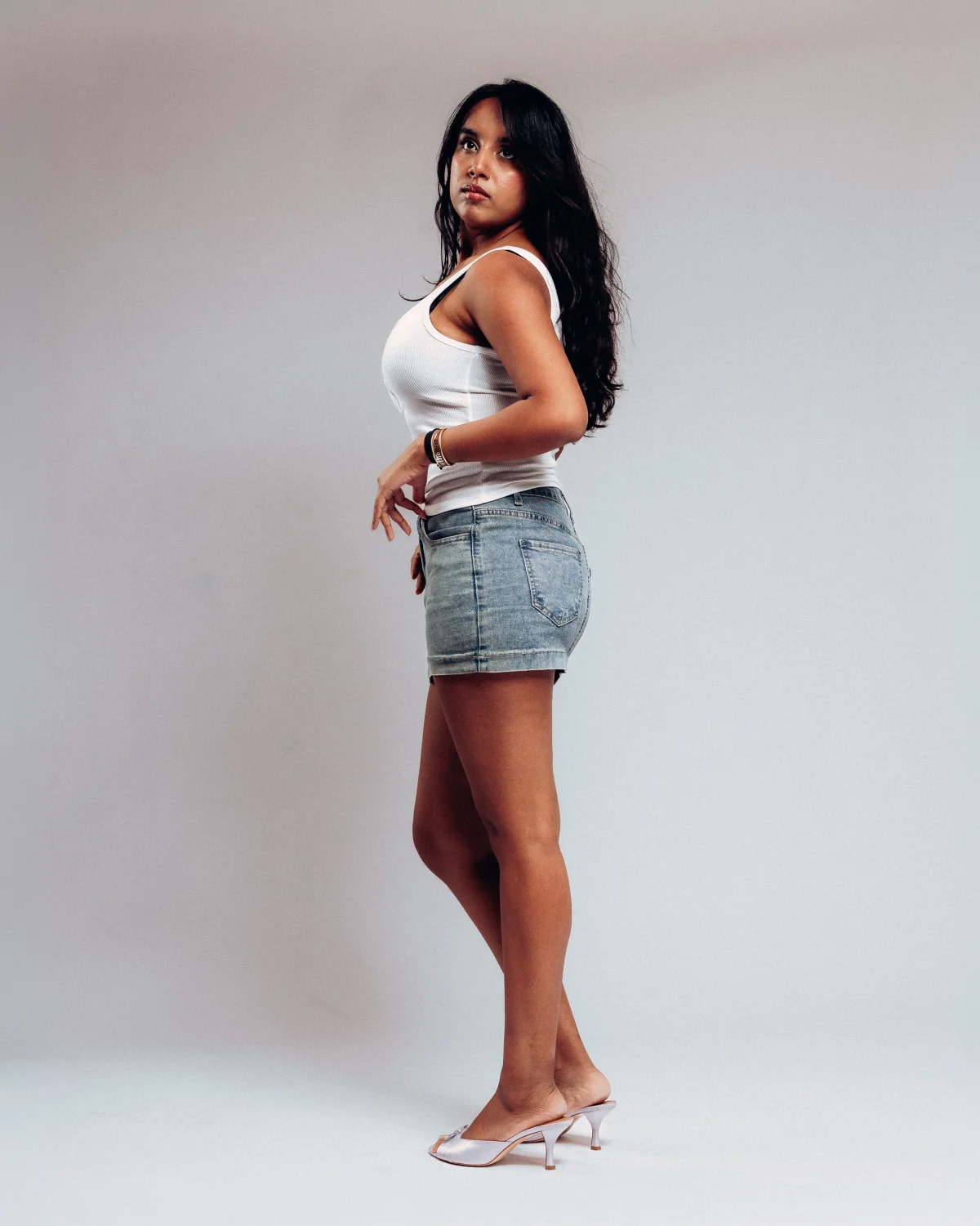 Woman with long dark hair posing in a white tank top and denim mini skirt against a plain grey studio background.